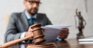 A bearded man with black glasses, grey blazer, white shirt and blue tie is sitting at a wooden desk reviewing papers with a gavel and statue of justice next to him.