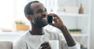 A side profile of a man at home wearing a white long-sleeved shirt holding a mug and talking on his cell phone with a smile on his face