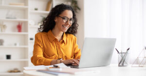 A curly haired woman with glasses and a yellow tee shirt sits at a white desk smiling and reading on her laptop