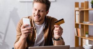 Smiling man in a tan shirt is paying his medical bill on his phone with his credit card in his other hand, sitting in his home with his laptop open in front of him