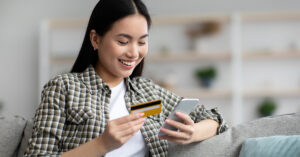 Young woman sitting on the couch wearing a flannel shirt holding her cell phone and credit card