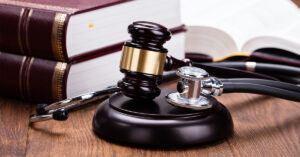 A gavel and stethoscope lay next to each other near three brown and gold law books on a wooden table