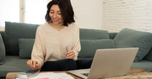 A woman with dark hair and a beige sweater is smiling on a grey couch working on paperwork with a calculator and laptop next to her on a table.