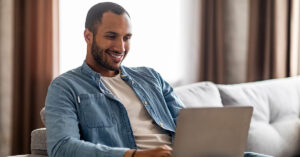 A short hair, bearded man wearing a blue button down shirt is sitting on a white couch in his home while smiling and using his laptop