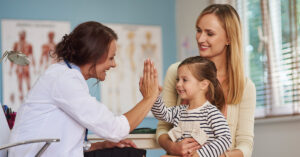 A child sitting in her mothers lap, high fiving her doctor in the doctors office