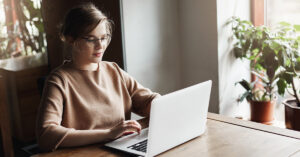 A woman with glasses sitting at a table in her home is working on her laptop