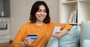 Woman with short dark hair wearing a mustard yellow top is sitting on a couch in her home making a credit card payment on her phone