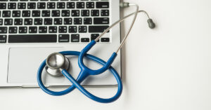 A silver laptop with black keyboard keys is on a white table with a blue stethoscope on the laptop