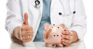 A doctor with a stethoscope around their neck is gesturing a thumbs-up with one hand and their other hand is resting on a pink piggy bank that has a band aid on it