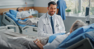 A male doctor is sitting next to an elderly patient who is laying on a hospital bed.