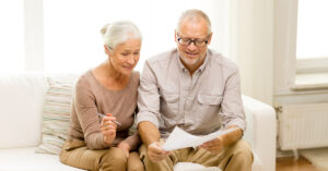 A senior couple wearing neutral colors are sitting on a couch and reviewing paperwork