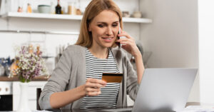 A woman is sitting in her home at the kitchen table while on the phone looking at her credit card and her laptop in front of her on the table