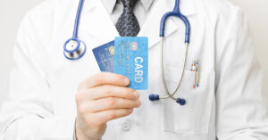 A doctor in a white coat has a blue stethoscope around his neck and is holding two blue credit cards in his hand