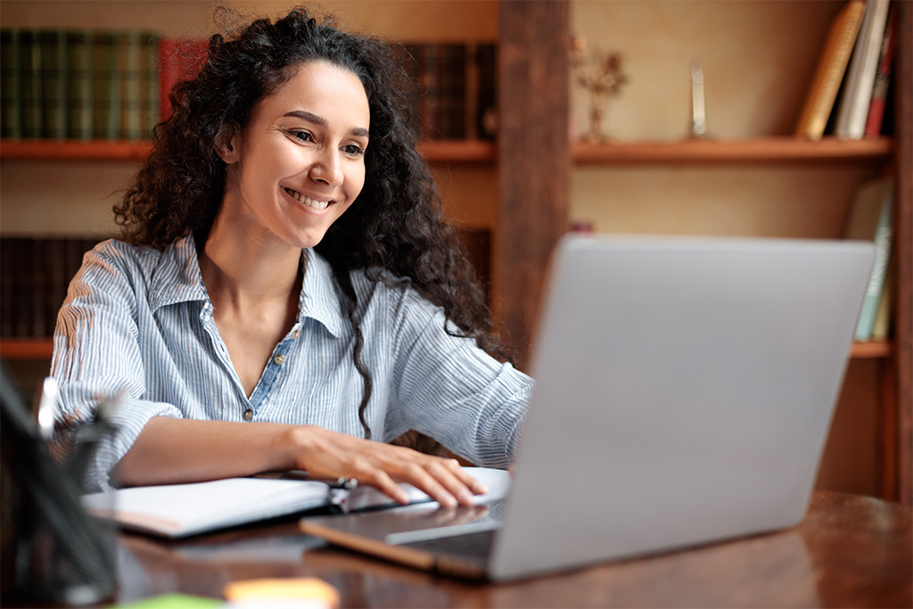 Woman with curly hair is smiling and working on her silver laptop