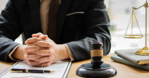 A man in a suit is sitting at his desk with his fingers interlocked atop a clipboard with a pen, a gavel next to him