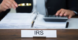 An office name plate says "IRS" with a person sitting at a desk holding a magnifying glass and auditing papers