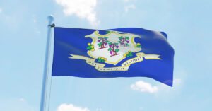 The blue Connecticut state flag on a flagpole and waves in the wind in front of a partly cloudy blue sky