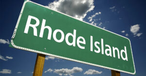 A green and white road sign that reads "Rhode Island" with a blue sky and clouds behind it