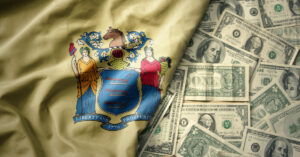 The New Jersey State flag is laying atop scattered $1- and $10- and 100-dollar bills