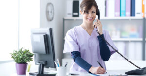 A medical receptionist is smiling on the phone and writing notes on a clipboard