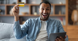 A man is sitting on a couch in his home holding a credit card and tablet and smiling