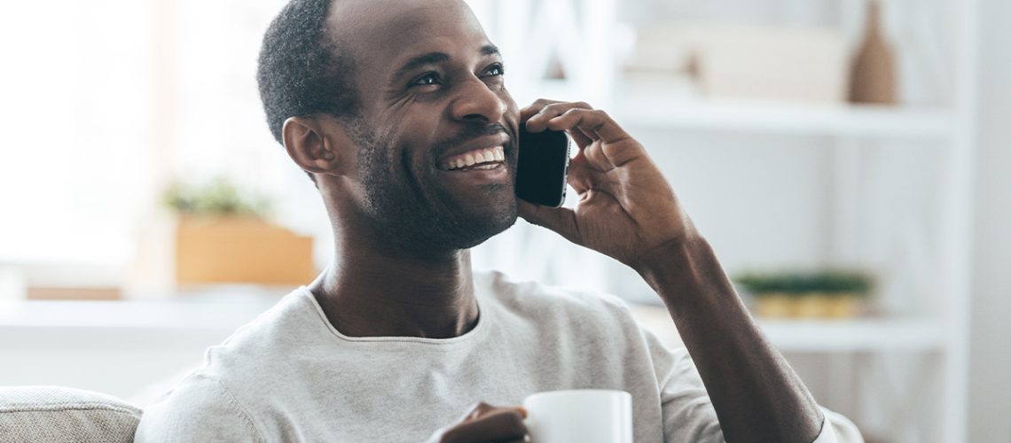 A side profile of a man at home wearing a white long-sleeved shirt holding a mug and talking on his cell phone with a smile on his face