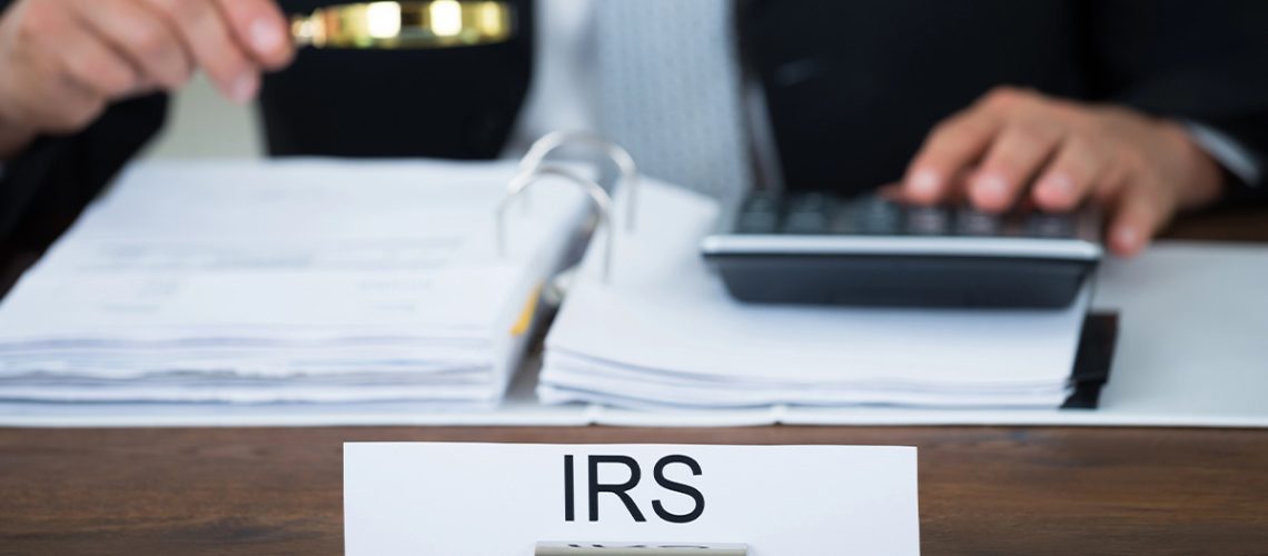 An office name plate says "IRS" with a person sitting at a desk holding a magnifying glass and auditing papers