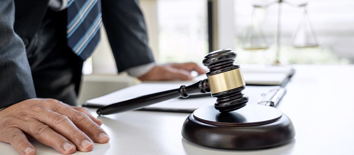 A man wearing a striped tie leans on his desk with papers and a gavel