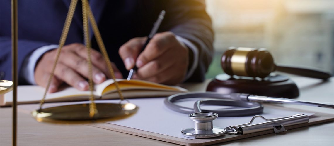 A person in a suit is writing on a notebook with a gavel, scale of justice, clipboard, and stethoscope on the table.