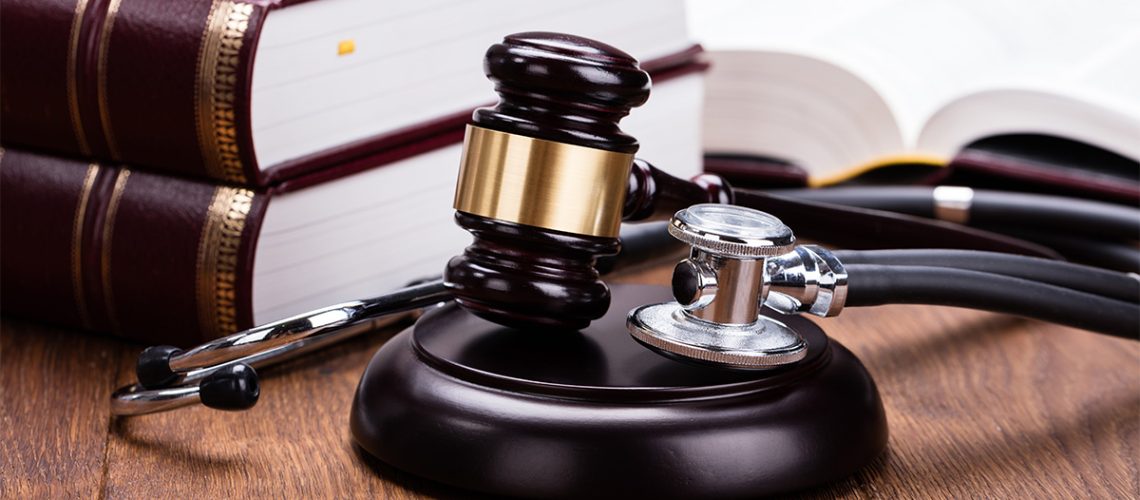 A gavel and stethoscope lay next to each other near three brown and gold law books on a wooden table