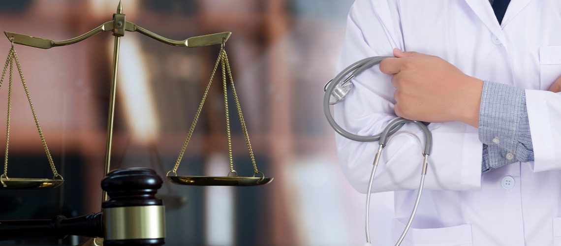 A doctor is holding a stethoscope and standing next to the scale of justice and a gavel