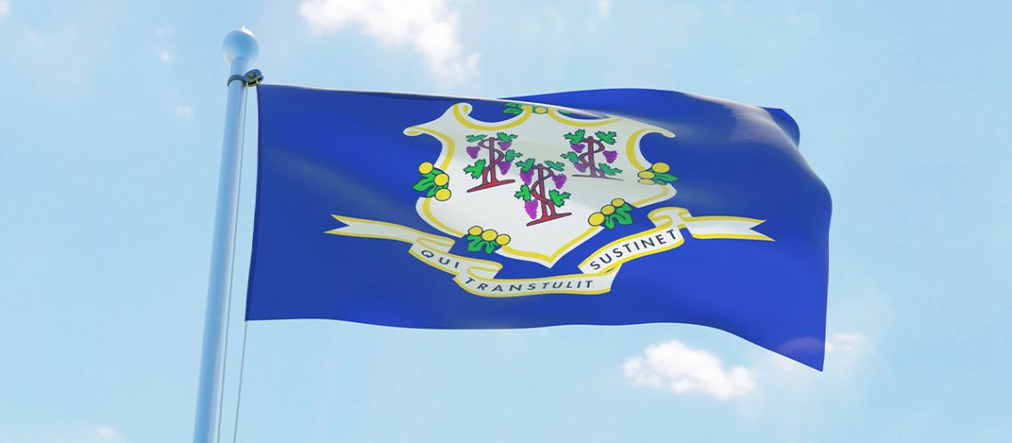 The blue Connecticut state flag on a flagpole and waves in the wind in front of a partly cloudy blue sky