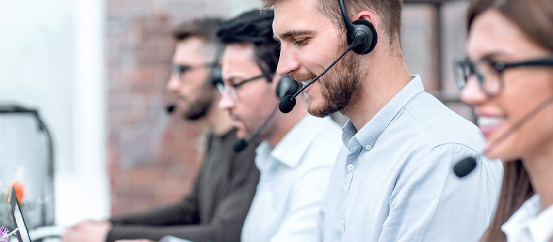 Three male and one female call center workers smiling, sitting in a line with headsets on and typing on their computers.