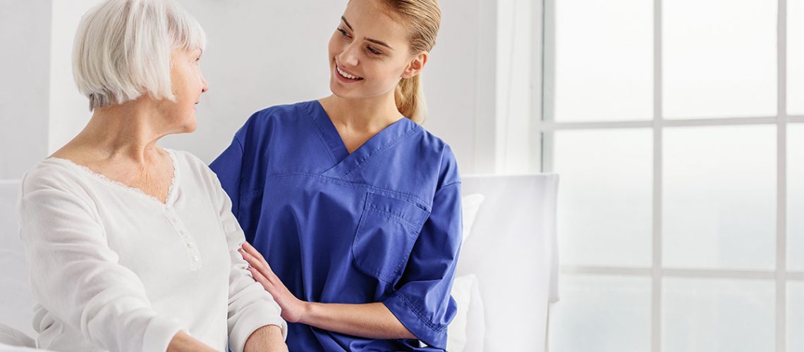 Blonde female nurse assisting a senior female patient in a white room with windows