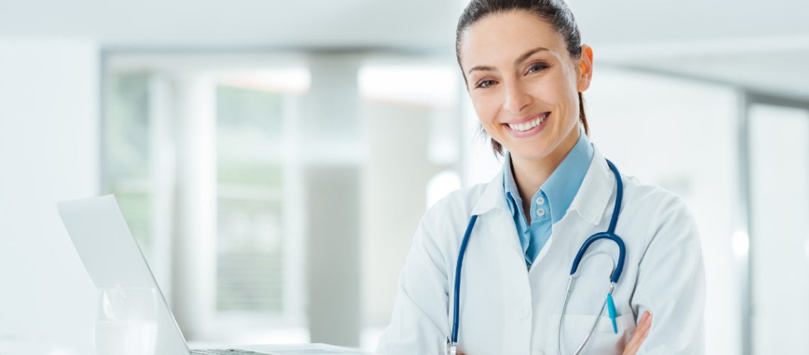 Female doctor with white lab coat and a blue stethoscope around her neck has her arms crossed and standing next to a table with an open laptop on it