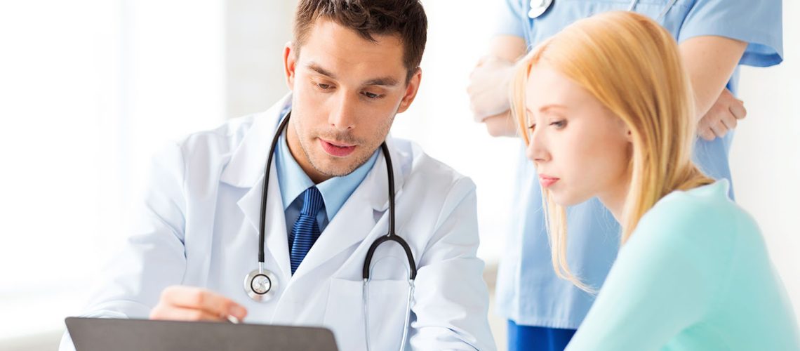 male doctor sitting next to a blonde female patient looking at a clipboard discussing information wiht a female doctor standing behind them listening in on the discussion.