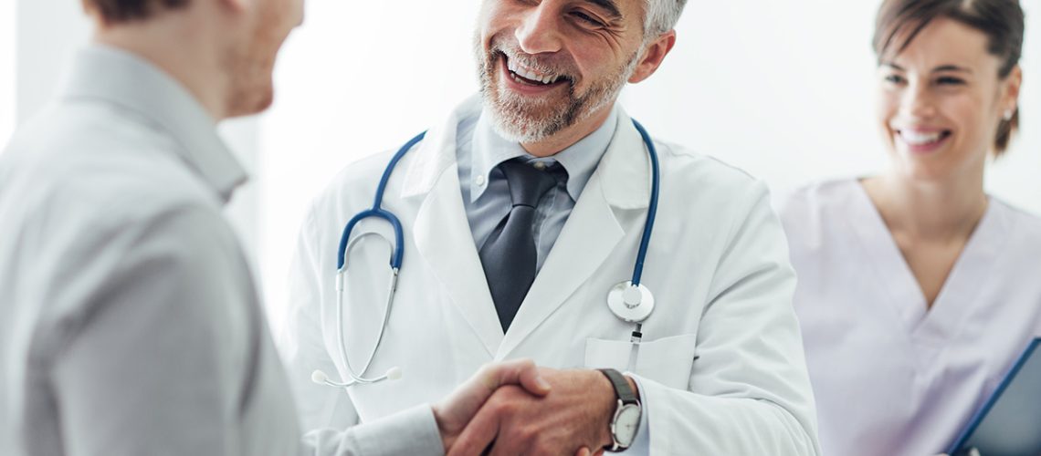 Male doctor wearing a white coat, tie, and stethoscope shaking a male patient's hand, both are smiling.