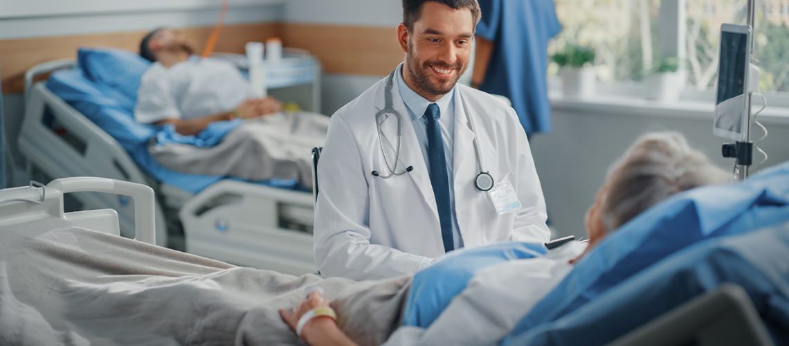 A male doctor is sitting next to an elderly patient who is laying on a hospital bed.