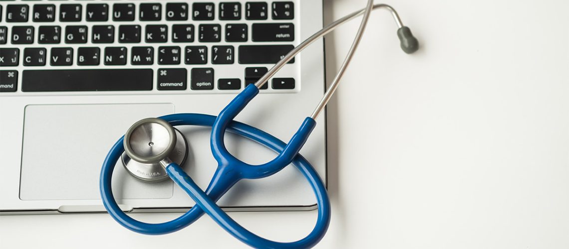 A silver laptop with black keyboard keys is on a white table with a blue stethoscope on the laptop