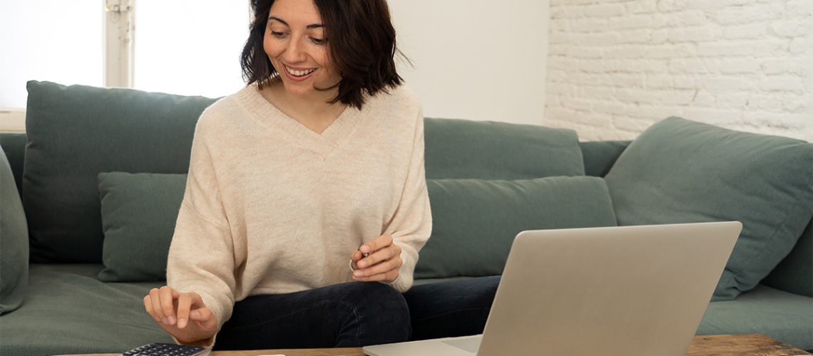 A woman with dark hair and a beige sweater is smiling on a grey couch working on paperwork with a calculator and laptop next to her on a table.