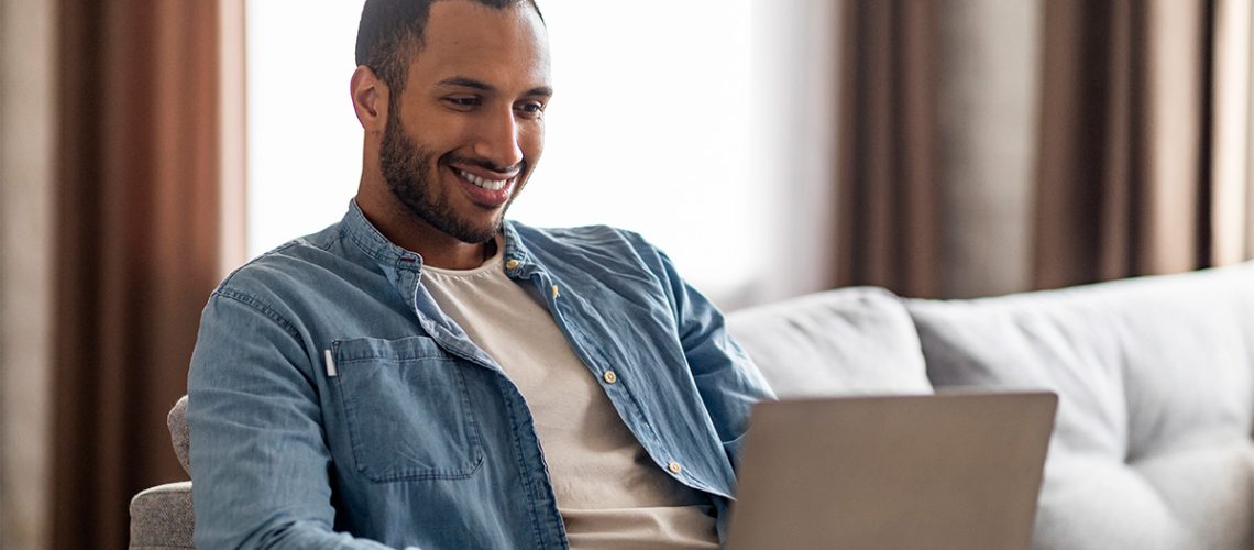 A short hair, bearded man wearing a blue button down shirt is sitting on a white couch in his home while smiling and using his laptop