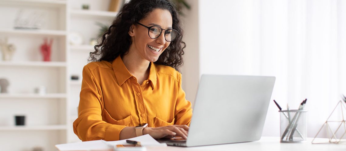 A curly haired woman with glasses and a yellow tee shirt sits at a white desk smiling and reading on her laptop