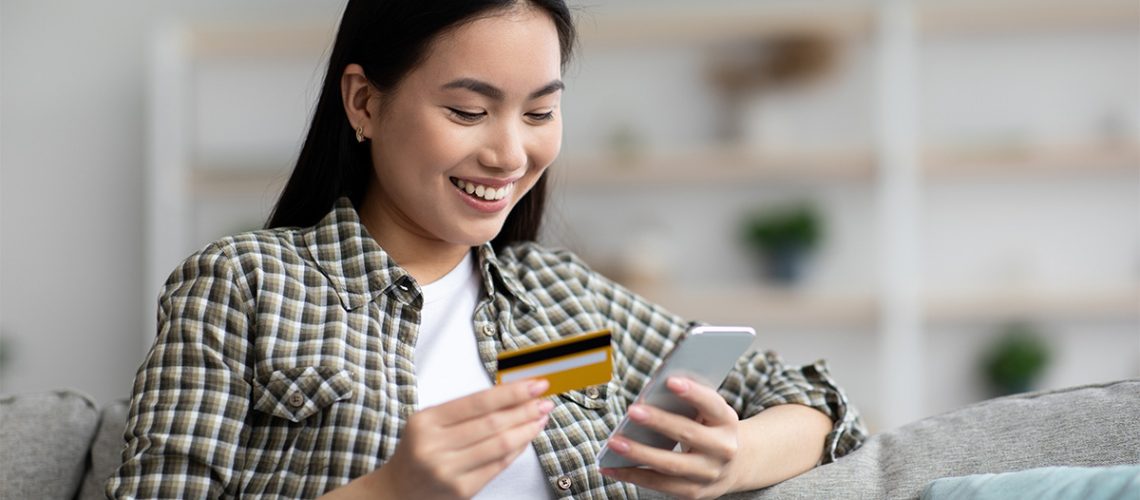 Young woman sitting on the couch wearing a flannel shirt holding her cell phone and credit card