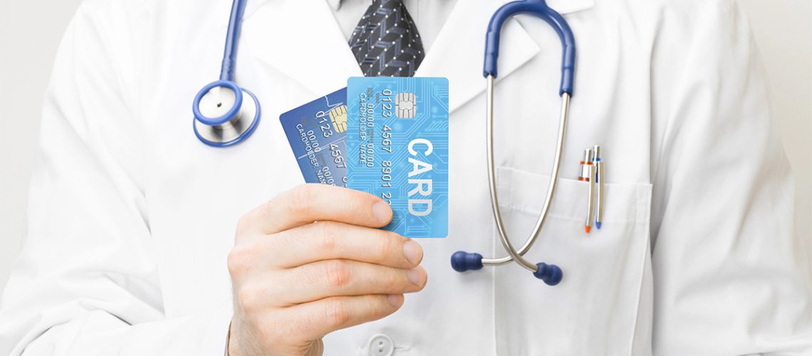 A doctor in a white coat has a blue stethoscope around his neck and is holding two blue credit cards in his hand