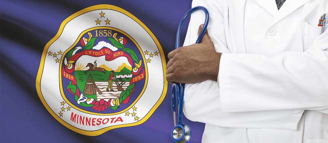 A doctor with a white coat and blue stethoscope stands in front of the Minnesota state flag