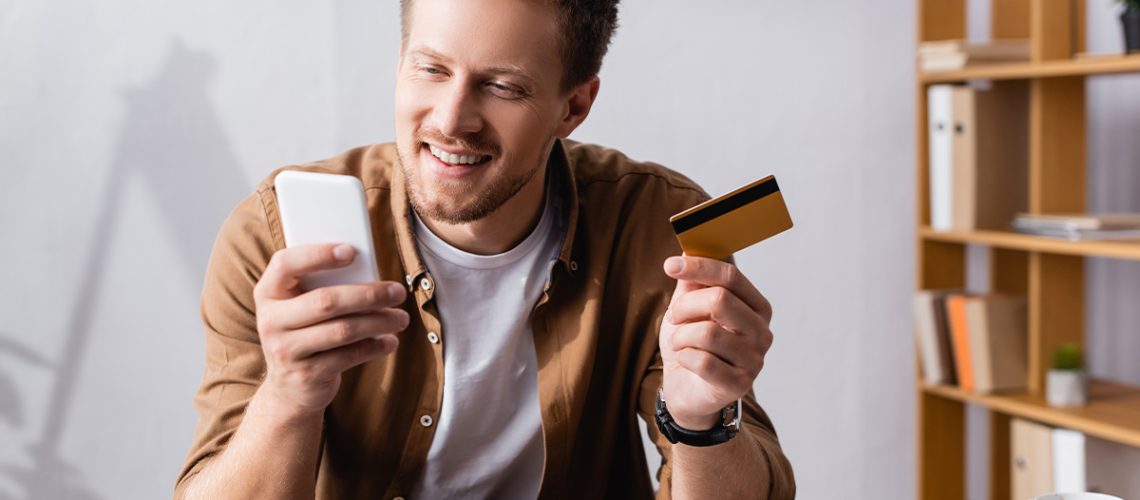 Smiling man in a tan shirt is paying his medical bill on his phone with his credit card in his other hand, sitting in his home with his laptop open in front of him