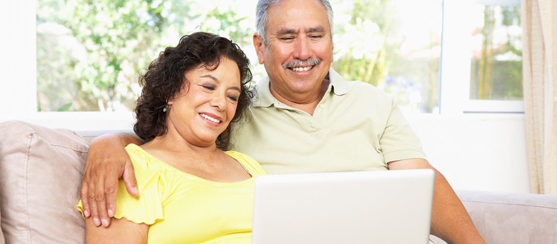 A smiling senior couple sitting on a couch in their home using a laptop