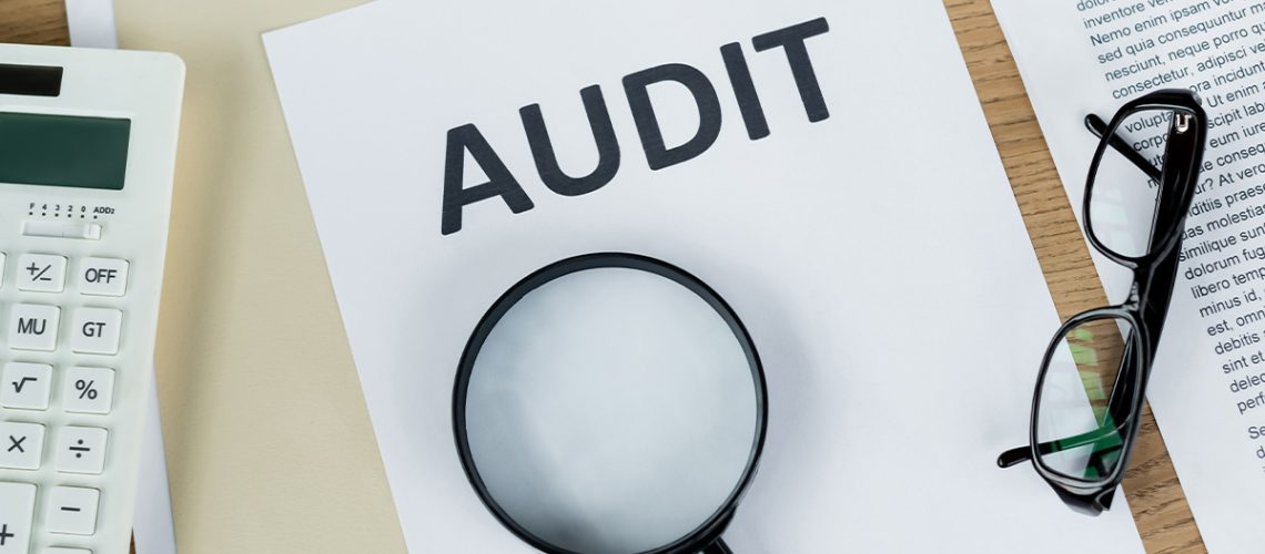 A folder, paper that reads "AUDIT", another piece of paper out of frame that has various text on it, a white calculator, a black pair of reading glasses, and a black magnifying glass, all on top of a wooden table.