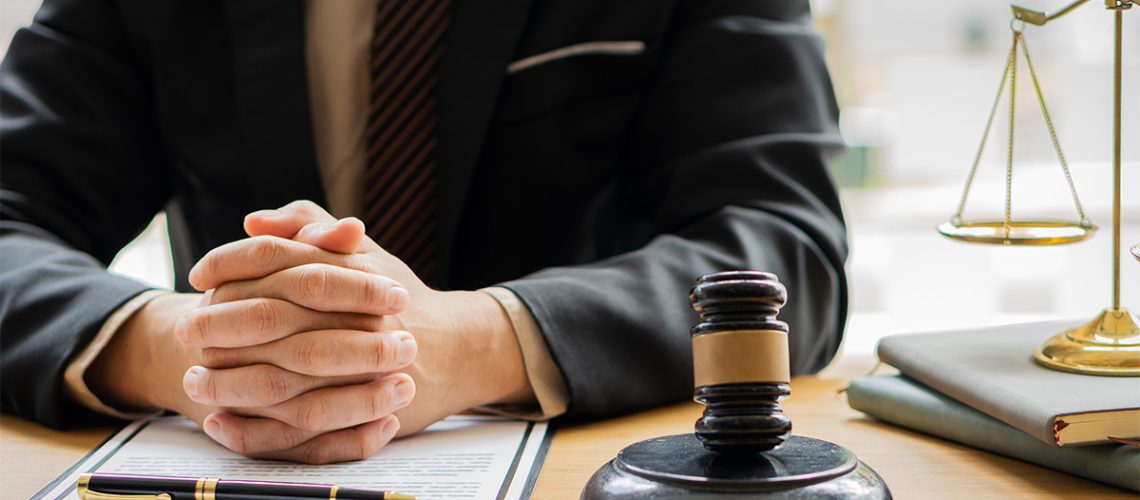 A man in a suit is sitting at his desk with his fingers interlocked atop a clipboard with a pen, a gavel next to him