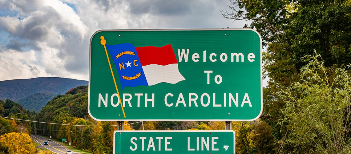 A road sign that says "Welcome To North Carolina State Line" with a landscape behind it